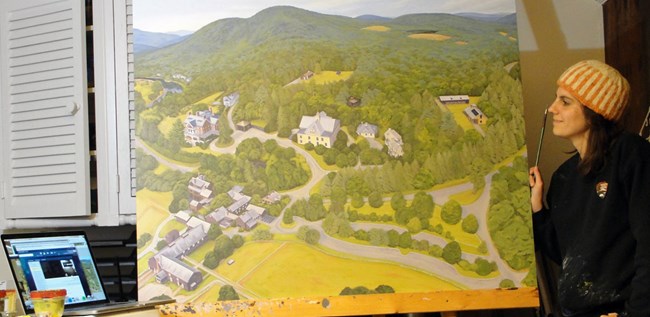 A woman stands next to a large painting of an aerial view of a developed area at the base of a mountain.